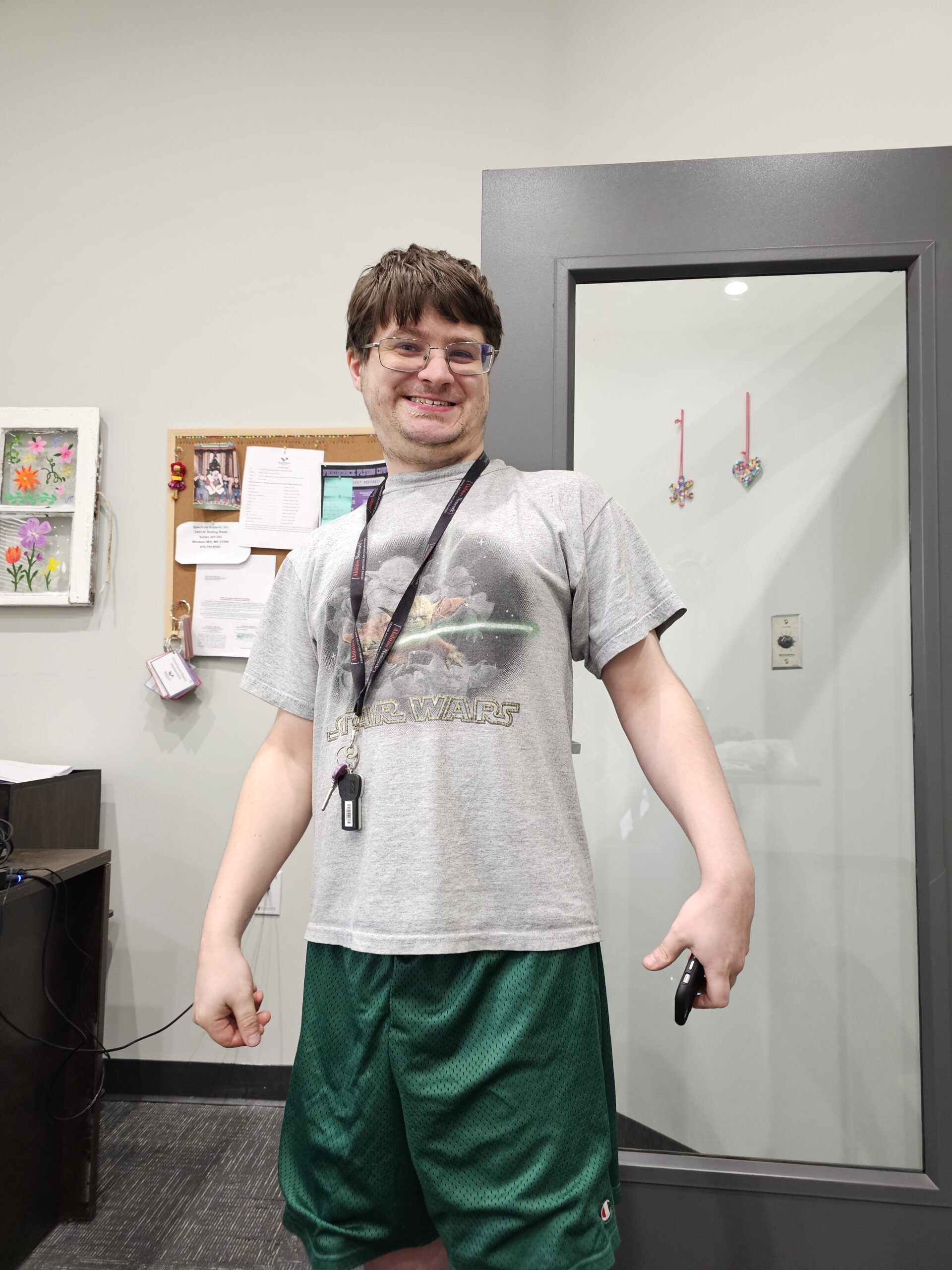 man stands smiling in office wearing white graphic tee shirt and green shorts