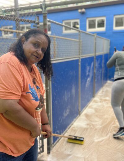 woman in orange shirt holding a mop and mopping outdoor corridor, with another woman in the background also mopping
