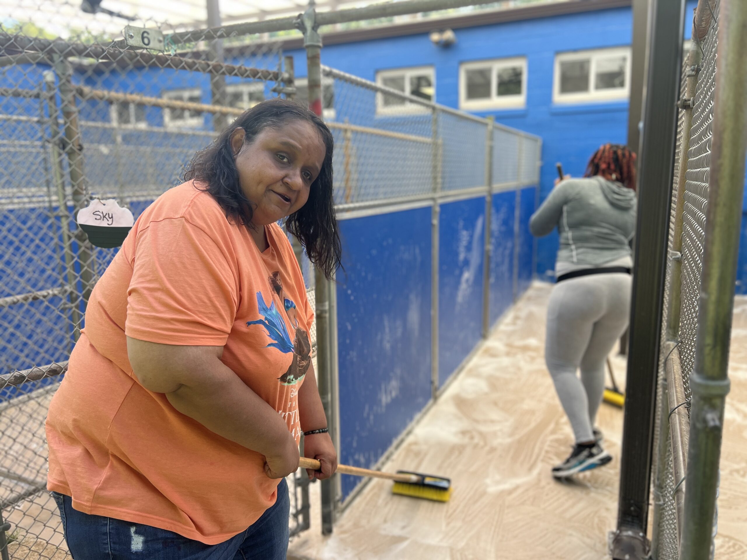 woman in orange shirt holding a mop and mopping outdoor corridor, with another woman in the background also mopping